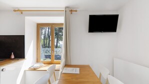 A room with a wooden table, white chairs, a TV, a window with curtains, and a view of trees. - Lis Martagon Mountain Cottage, 3-Star Comfort Near Lifts (Font-Romeu-Odeillo-Via)