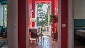A room with a red wall, a wooden desk, a chair, a lamp, a potted plant, and a view of trees through a window. - Bohémien Rome (Roma)