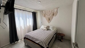 Ein Schlafzimmer mit einem Bett, einem an der Wand montierten Fernseher, einem Wandbehang aus Makramee und einem kleinen Nachttisch.