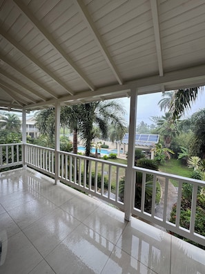 A covered balcony with a tiled floor and white railings, overlooking a pool and lush greenery.