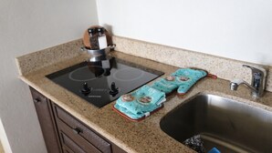 Una cocina con tope de inducción negro, un lavamanos de acero inoxidable y dos toallas de cocina verde aguamarina con motivos tropicales.