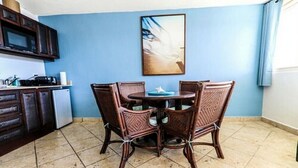 Un comedor con una mesa redonda y cuatro sillas, una cocina con armarios oscuros y un microondas, y una pared azul con un cuadro enmarcado.