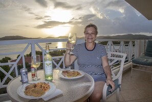 A person seated at a table with a plate of food, a glass of wine, and a bottle of Cuvée wine. The table is on an outdoor balcony with a view of a body of water and mountains in the background.