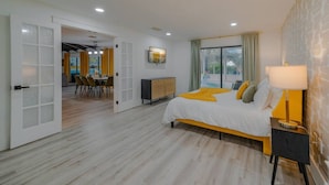 Et moderne hotelværelse med en stor seng, et natbord med en lampe, udsigt til det fri gennem et vindue, og et spiseområde, der kan ses gennem en åben dør.
