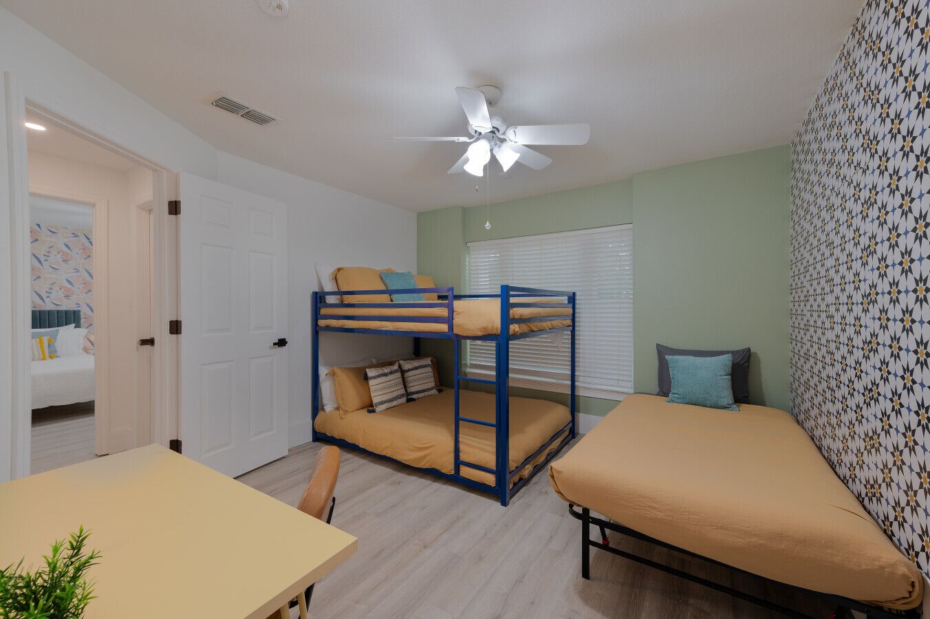 A bunk bed room with a ceiling fan, a table, and a window with blinds.