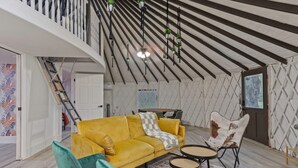 A modern loft with a high ceiling, exposed wooden beams, a yellow sofa, teal armchairs, and a patterned rug. - Florida Yurt Escape • Nature + Comfort (Kissimmee)