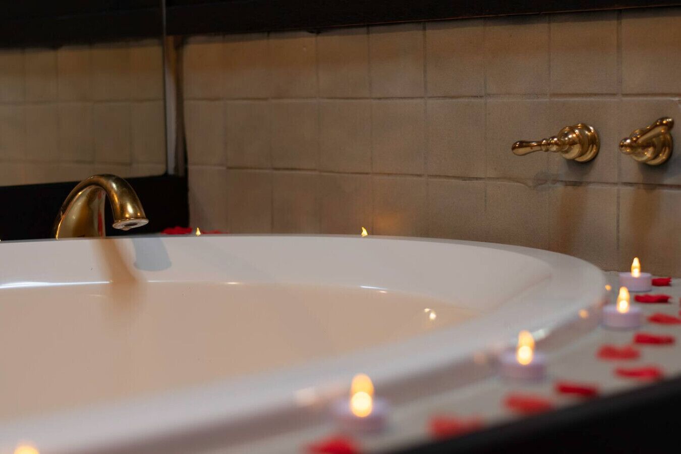 Eine weiße Badewanne mit goldfarbenem Wasserhahn und zwei goldenen Griffen, umgeben von Kerzen und Rosenblättern.