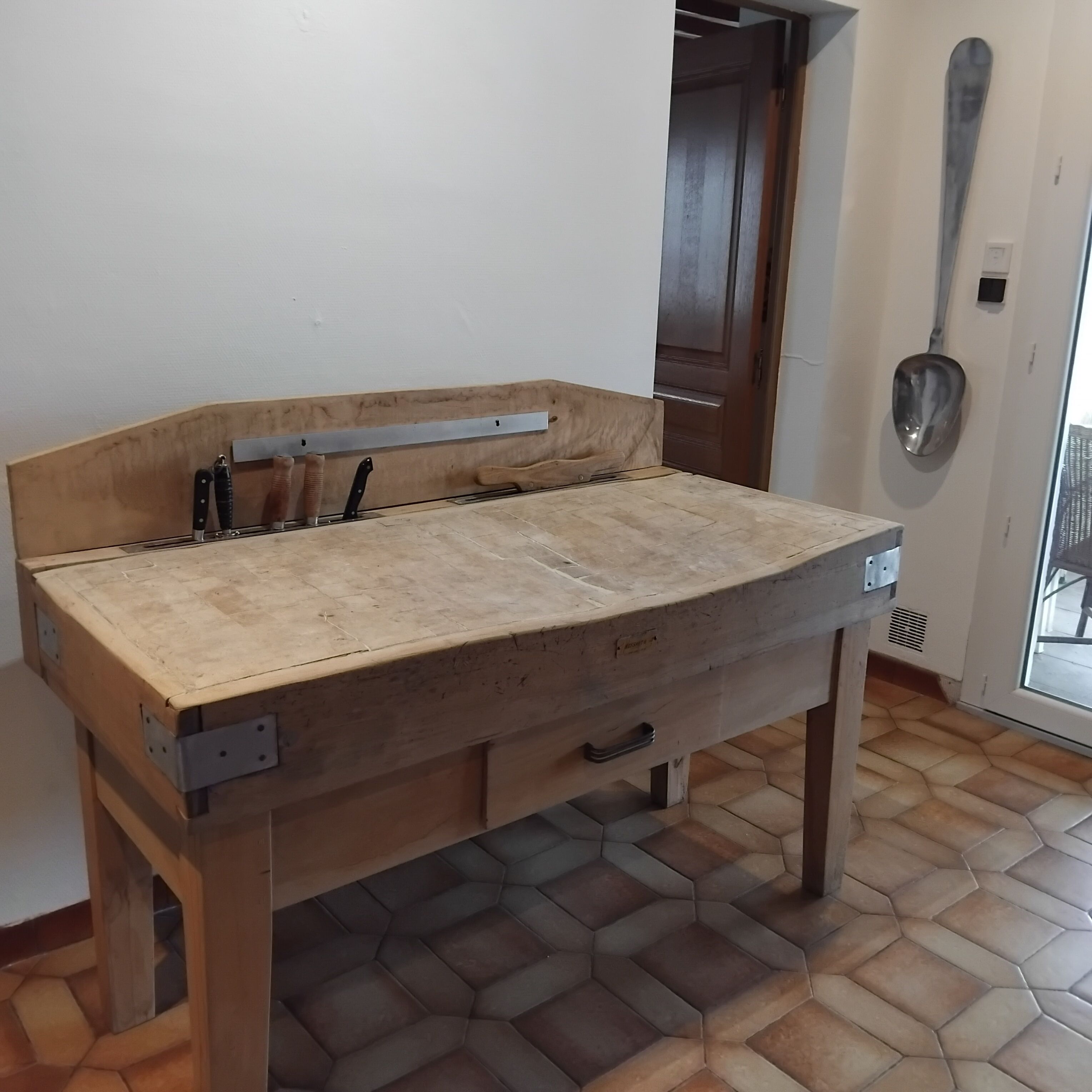 A wooden workbench with a metal shelf, a door in the background, and a metal spoon on the wall.
