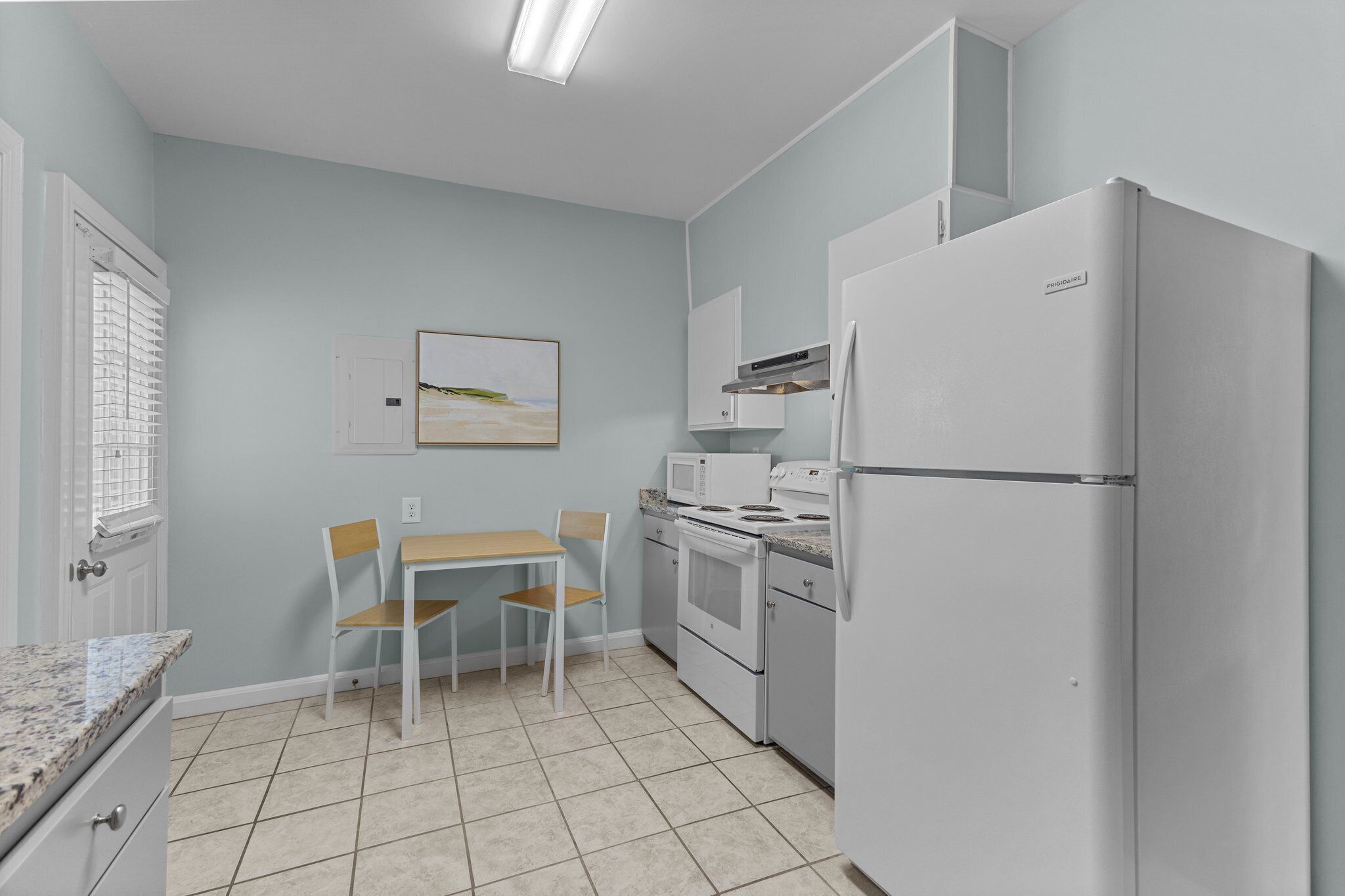Une cuisine de taille réduite avec des appareils électroménagers blancs, une table à manger avec des chaises et une fenêtre avec des stores.