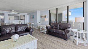 Una sala de estar moderna con dos sofás de cuero marrón, una mesa de centro blanca y una mesita auxiliar con cubierta de vidrio y una lámpara. Se observa una cocina al fondo con armarios blancos y un área de comedor.