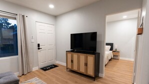 A modern, well-lit room with a wooden floor, a television on a cabinet, a bed with a nightstand, and a window with curtains.
