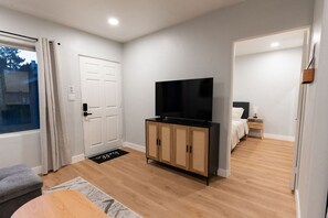 A modern, well-lit room with a wooden floor, a television on a cabinet, a bed with a nightstand, and a window with curtains. - Modern Hideaway in North Park - 3941 (San Diego)