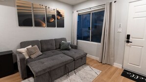 A modern living room with a grey sectional sofa, a large window with curtains, and a door with a keypad lock.