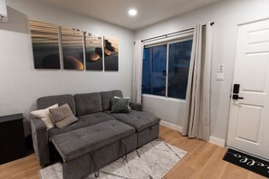 A modern living room with a grey sectional sofa, a large window with curtains, and a door with a keypad lock. - Modern Hideaway in North Park - 3941 (San Diego)