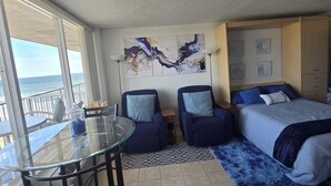 A hotel room with a balcony, a bed with blue bedding, a glass table with chairs, and a wall with abstract art. - Chic Cozy Condo with Stunning Views (Daytona Beach Shores)
