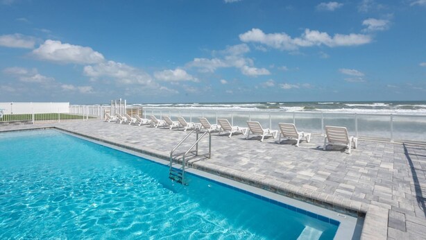 A swimming pool with lounge chairs facing the ocean, a clear sky, and a view of the beach. - Chic Cozy Condo with Stunning Views (Daytona Beach Shores)
