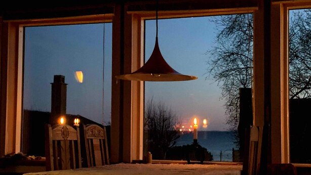 Een kamer met een eettafel, grote ramen, een hanglamp en een open haard.