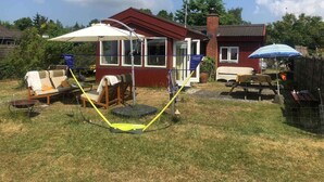 Een huis met een rode gevel, een overdekte veranda en een omheinde achtertuin. Er zijn buiten zitplekken met parasols en een picknicktafel.