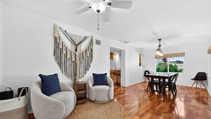 A spacious living room with a wooden floor, two armchairs, a dining table with chairs, a ceiling fan, and a large window with a view of greenery. - 3 miles to Siesta Key, Screened in Lanai, 3 Bedrooms, Walking dist to Publix (Sarasota)