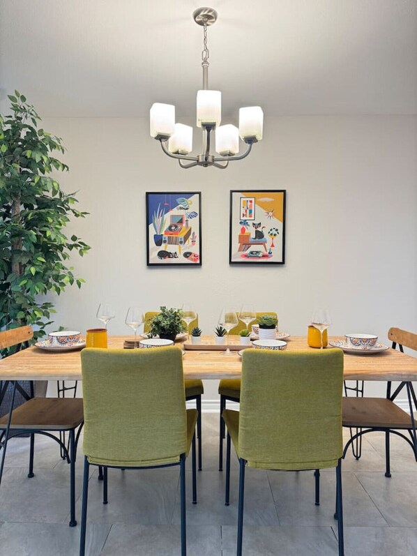 A dining area with a wooden table, four chairs, a chandelier, two framed artworks, and a potted plant.
