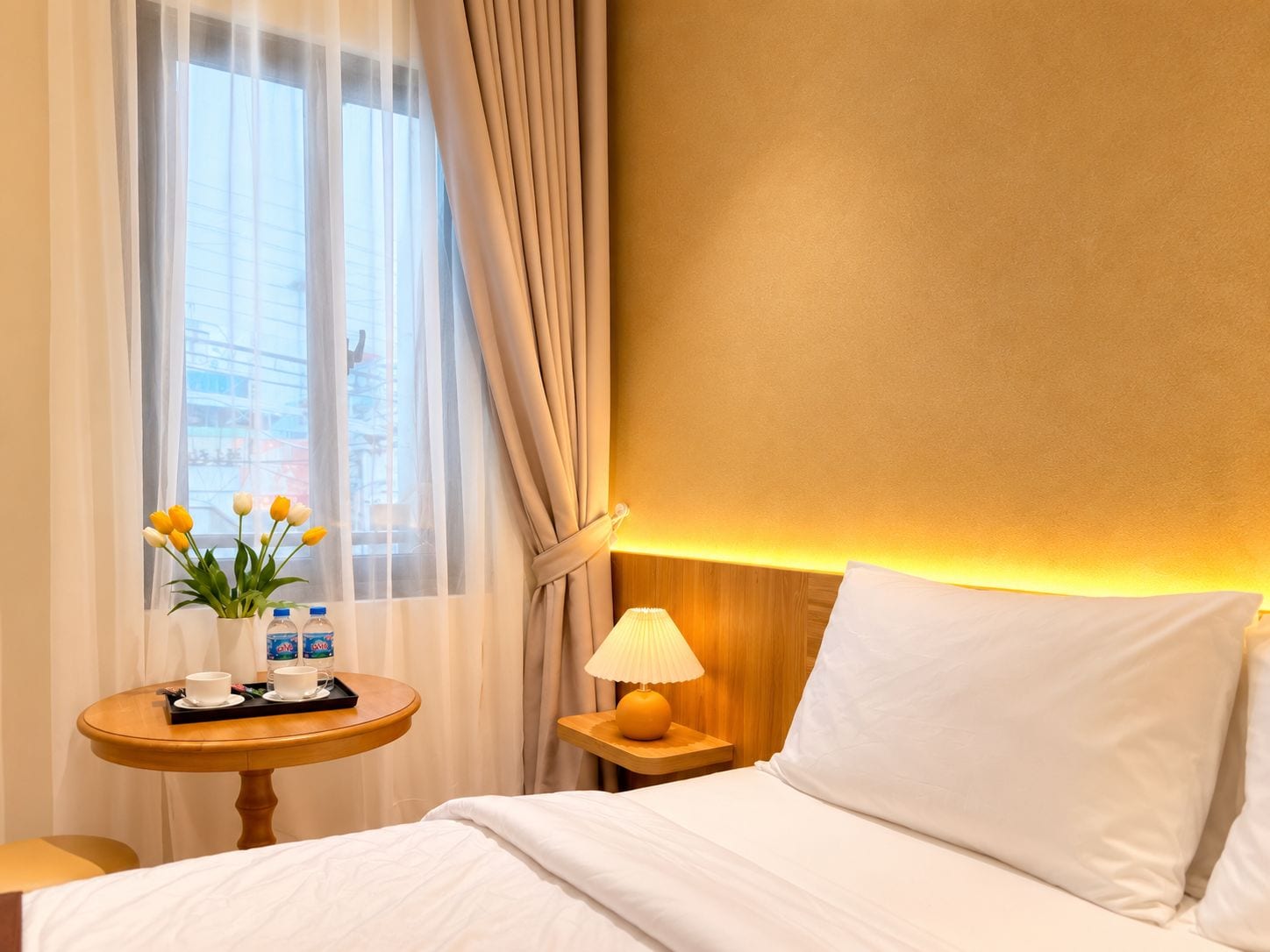 Een hotelkamer met een bed, een nachtkastje met een lamp, een kleine tafel met een bloevas en een dienblad met bekers en flessen.