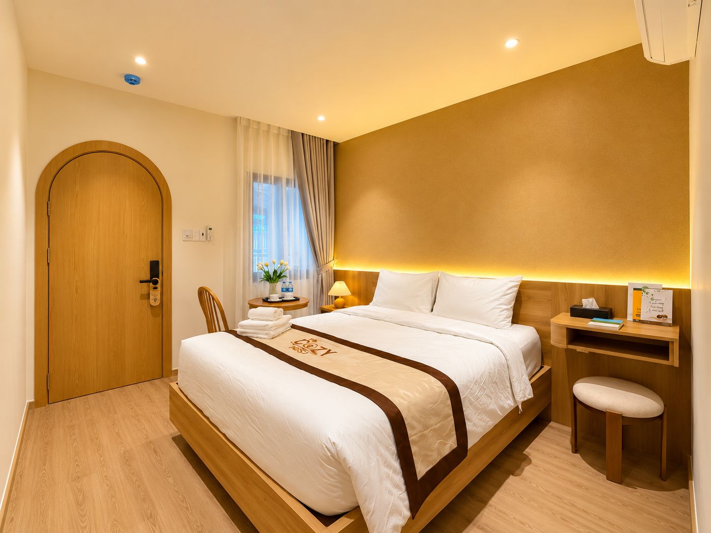 A hotel room with a wooden door, a bed with white bedding, a bedside table with a lamp, and a small stool.