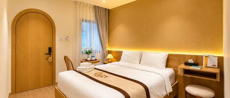 A hotel room with a wooden door, a bed with white bedding, a bedside table with a lamp, and a small stool.