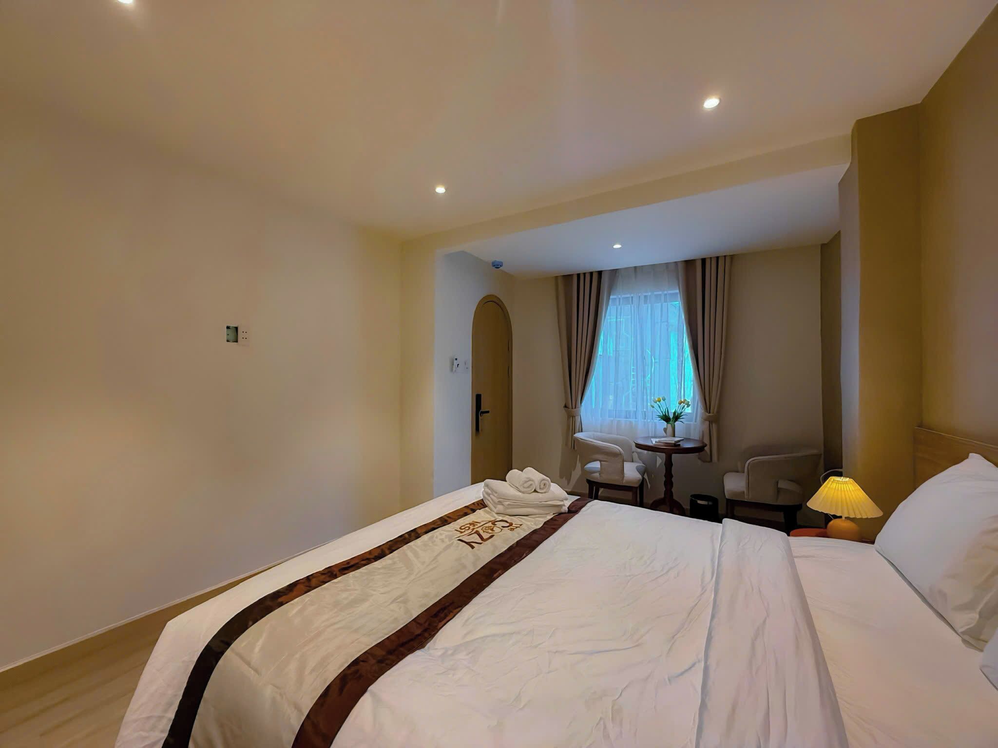 A hotel room with a large bed, a small table with chairs, a window with curtains, and a lamp on the bedside table.