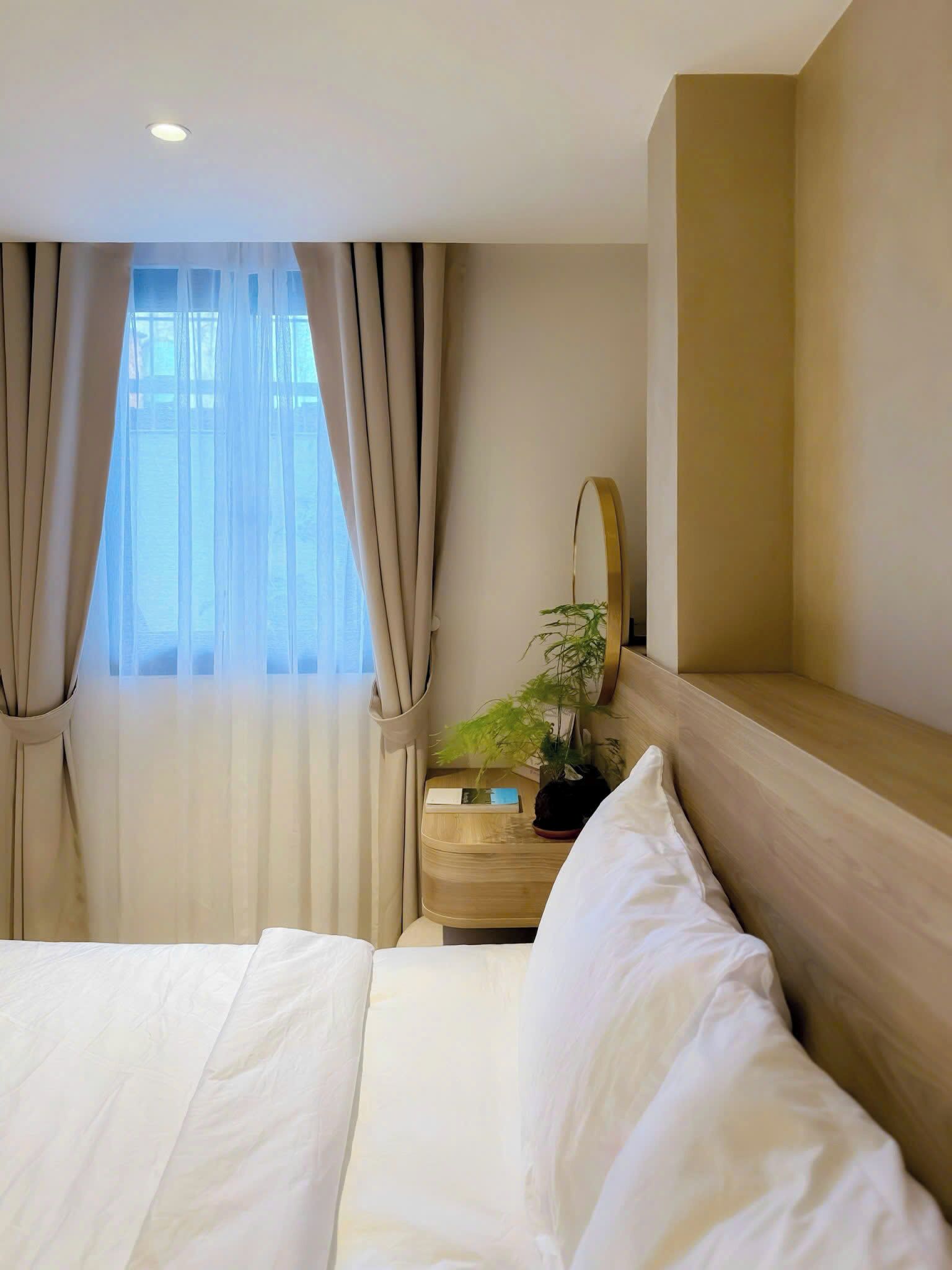 Quarto de hotel com uma cama, mesa de cabeceira com uma planta, espelho e janela com cortinas.