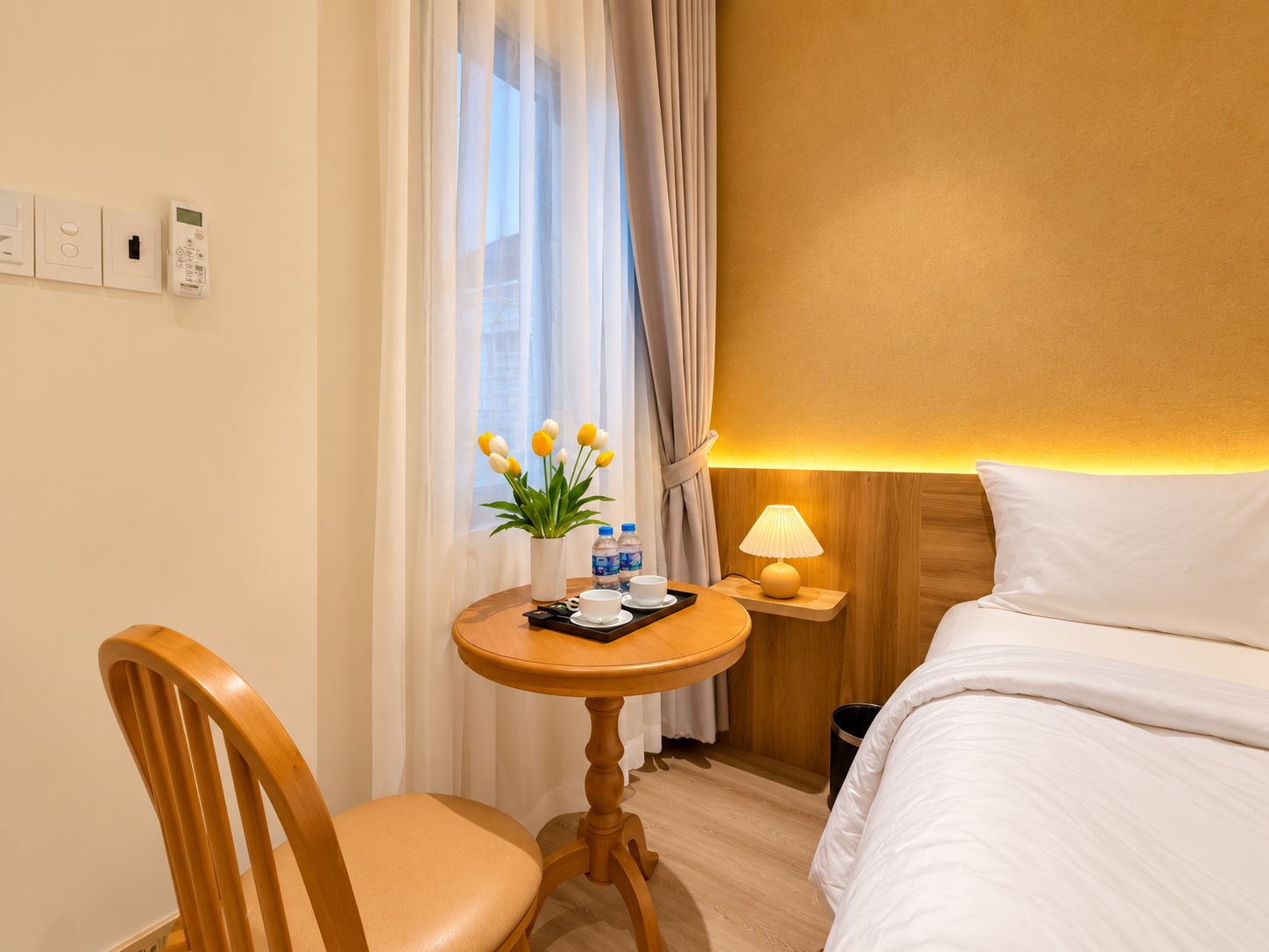 Habitación de hotel con cama, una silla de madera, una mesita redonda con una planta, una lámpara y un termostato en la pared.