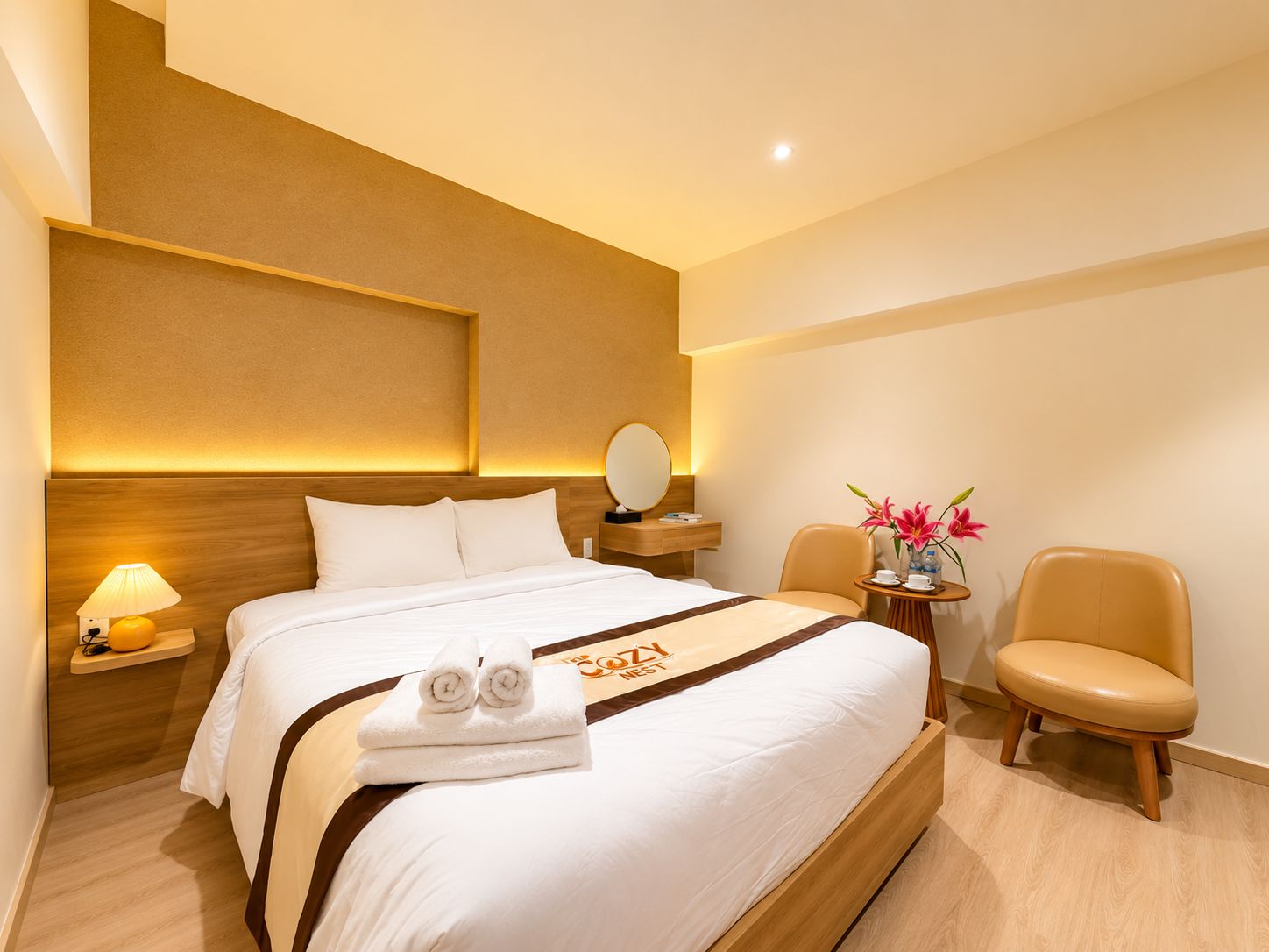 Een hotelkamer met een bed, nachtkastje, lamp, stoel en een tafeltje met bloemen.
