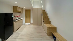 Une cuisine moderne avec un réfrigérateur noir, des meubles en bois et un petit comptoir agrémenté d’un vase de fleurs. Un escalier en bois mène à une porte située au fond du couloir.