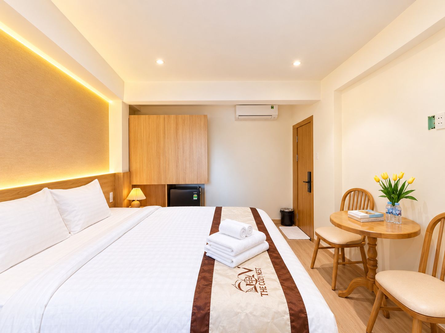 Ein Hotelzimmer mit einem ordentlich bezogenen Bett, einem kleinen Tisch mit Stühlen, einem Kühlschrank und einer Wand-Klimaanlage.