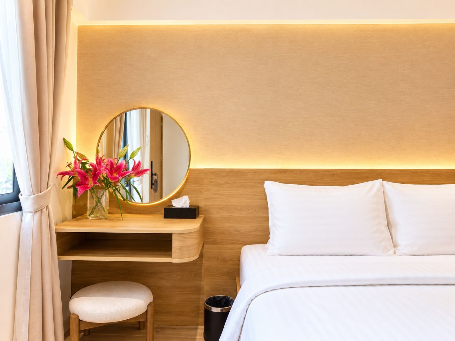 A hotel room with a bed, a bedside table with a mirror, a vase with flowers, and a small stool.