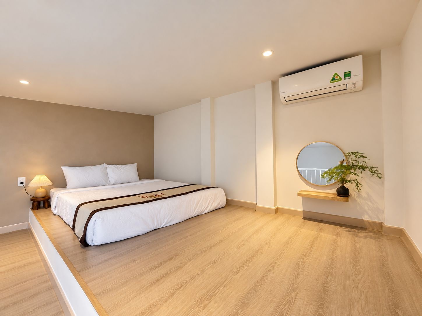 A modern hotel room with a bed, bedside lamp, air conditioning unit, and a small plant on a shelf.