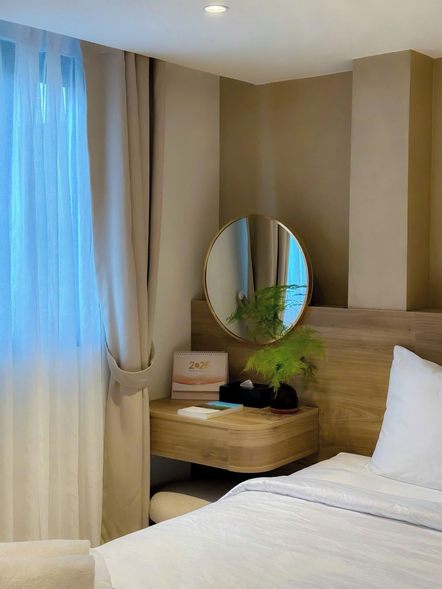 A hotel room with a bed, a bedside table with a mirror, a potted plant, and a calendar.