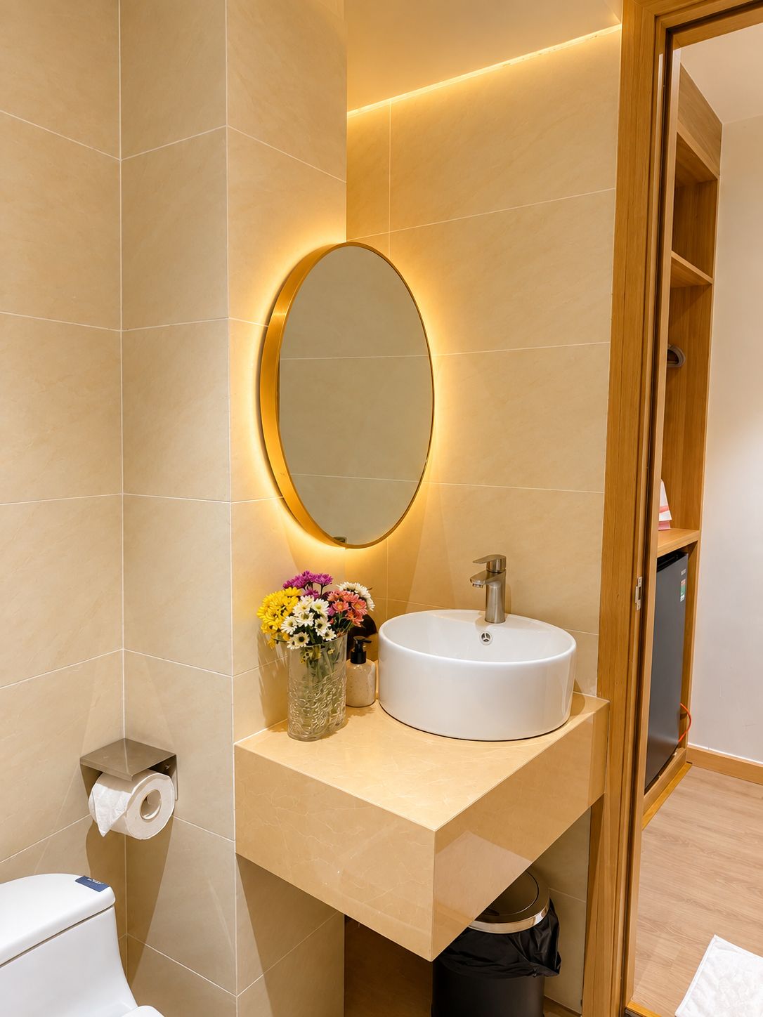A modern bathroom with a white sink, a round mirror with a light, and a vase of flowers.