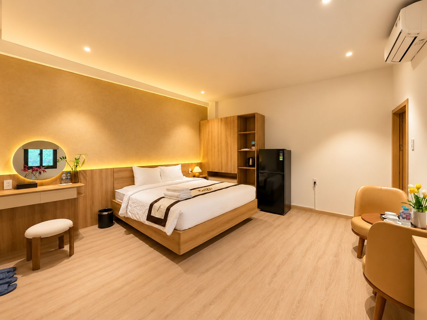 A modern hotel room with a bed, a desk with a computer, a refrigerator, and a seating area.