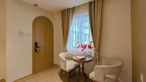 Une chambre d’hôtel avec une porte en bois, une petite table sur laquelle est posé un vase de fleurs rouges, deux chaises et une fenêtre avec des rideaux.