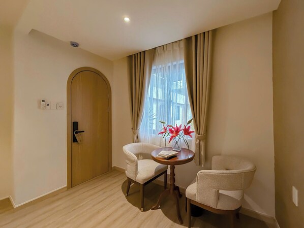 Habitación de hotel con puerta de madera, una mesita con un jarrón de flores rojas, dos sillas y una ventana con cortinas.