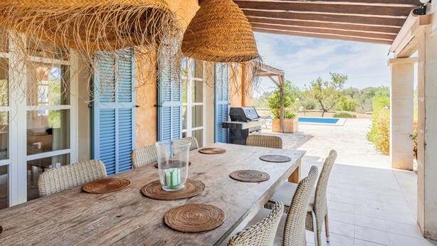 Ein Holztisch mit gewebten Tischsets und Korbstühlen, unter einem Strohdach mit Blick auf einen Pool und Grünflächen.