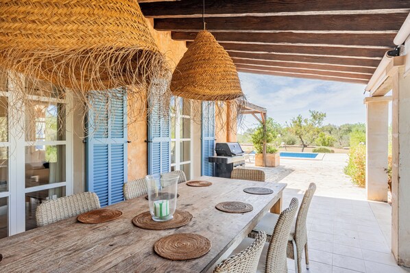 A wooden dining table with woven placemats and wicker chairs, under a thatched roof with a view of a pool and greenery. - Finca Los Almendros near Santanyi with private pool, wifi and air conditioning (Santanyi)