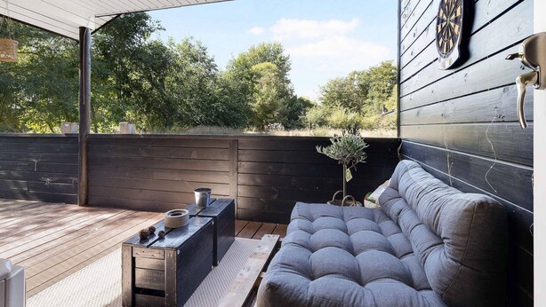 A covered outdoor seating area with a cushioned bench, a small table with a kettle, and a dartboard on the wall. - 5 Star Holiday Home in Sjaellands Odde (Sjællands Odde)