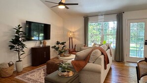 A spacious living room with a ceiling fan, a flat-screen TV, a sofa, a coffee table, and a large window with curtains. - Near Watts Bar | Pet-Friendly | 2 Kings | Easy Checkout (Rockwood)