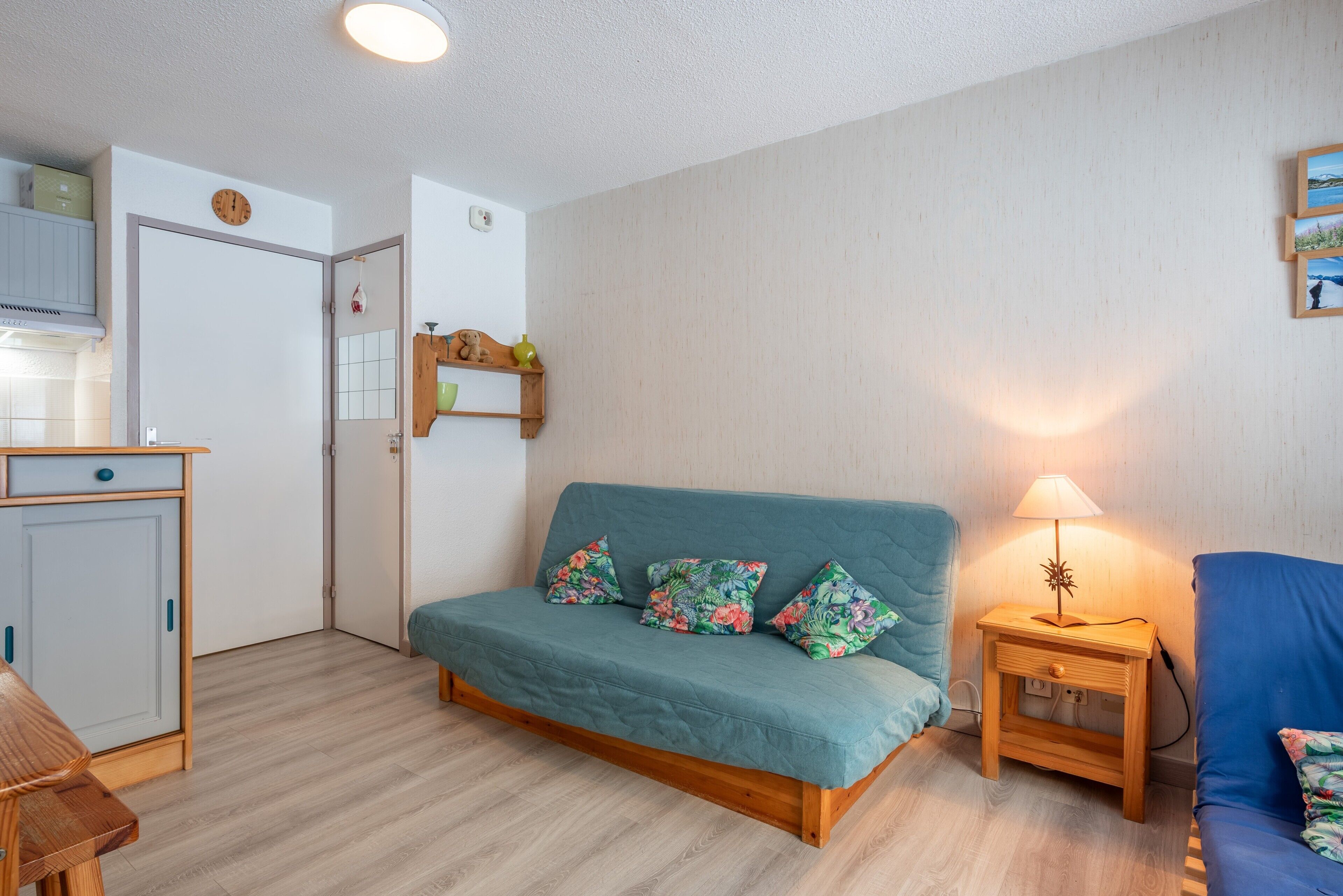 A room with a teal sofa, a wooden nightstand with a lamp, a wooden cabinet, and a door with a clock.