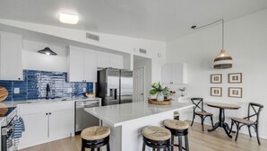 Une cuisine moderne avec des meubles blancs, un réfrigérateur en acier inoxydable, un pare-feu bleu et un îlot central doté de tabourets de bar.