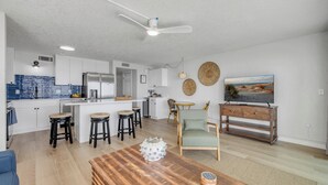 Une cuisine moderne avec des meubles blancs, un îlot central et des tabourets de bar. On y trouve également un coin repas avec une table et des chaises en bois, un téléviseur et une étagère fixée au mur.