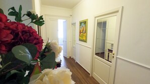 A hallway with white walls, wooden flooring, and a door with a glass panel. There is a painting on the wall and a vase with flowers on the left.