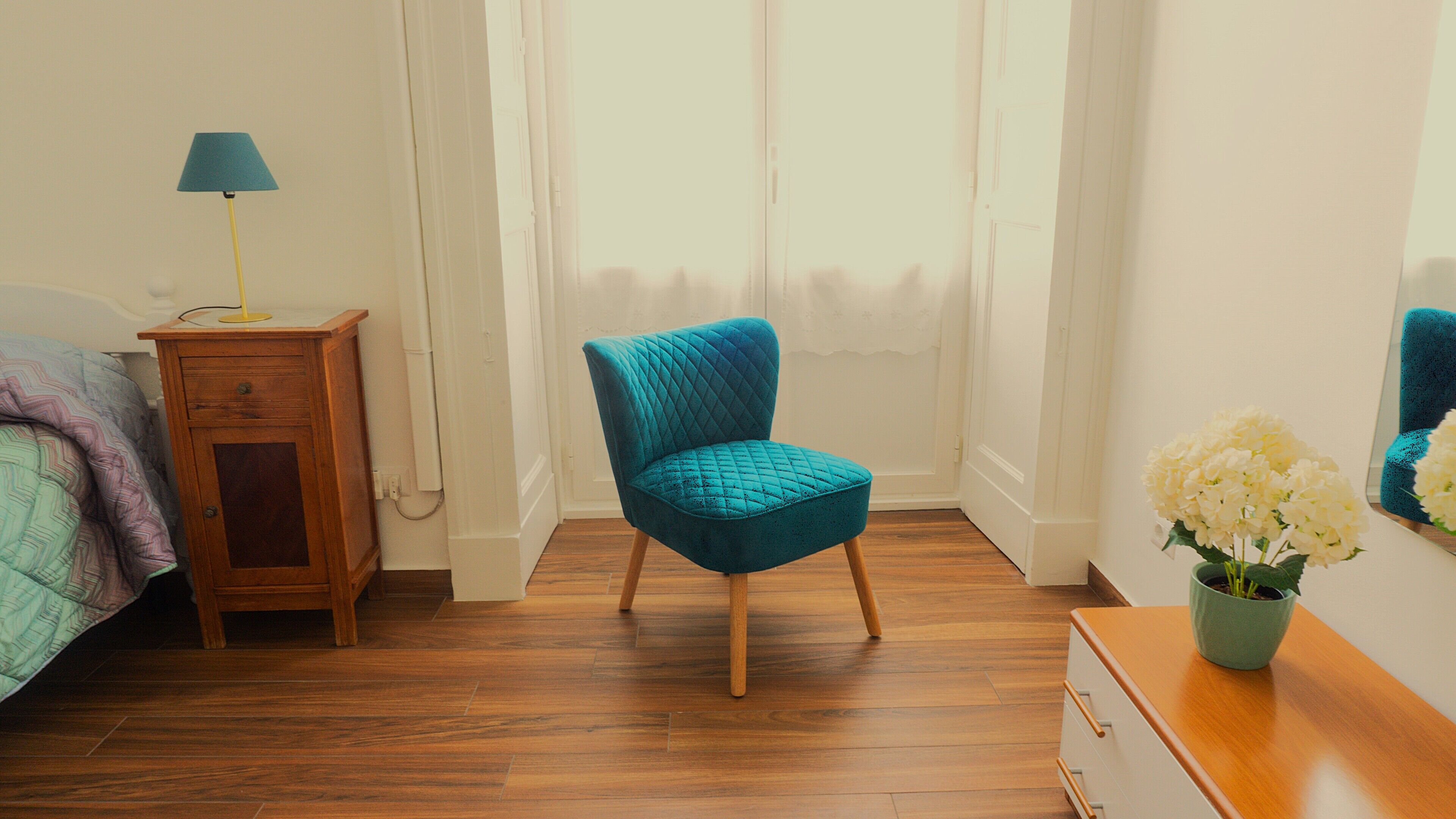 A bedroom with a teal upholstered chair, a wooden nightstand with a lamp, a dresser, and a mirror.