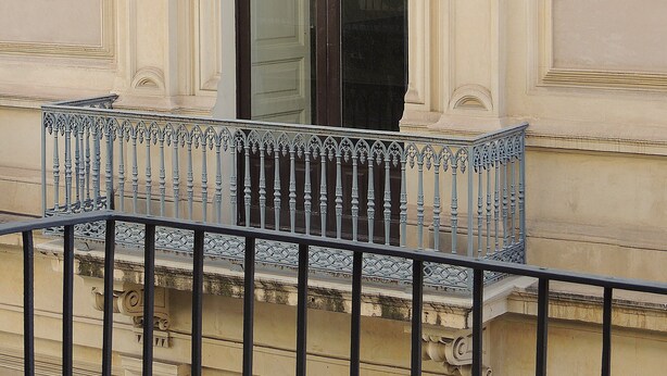 A building facade with an ornate arched window, a balcony with a metal railing, and classical architectural details.